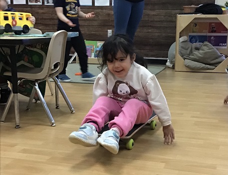 Preschool girl pushing self through room on skateboard with hands