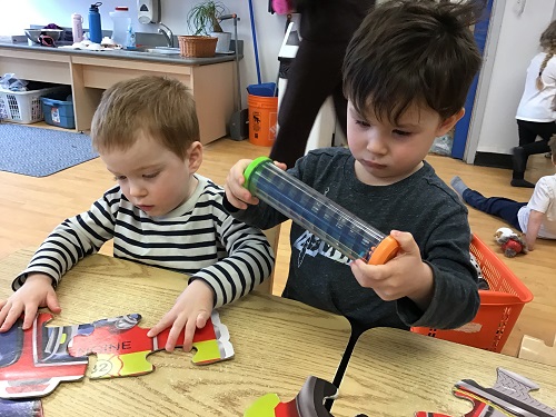 2 preschool children standing at the table, one child looking at a sensory tube and another putting together 2 puzzle pieces