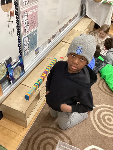 one school age child kneeling on the ground with rectangle small blocks lined up on some bigger blocks, child is looking at the camera with hands in his pocket. There are 3 other school age children laying on the ground in the background