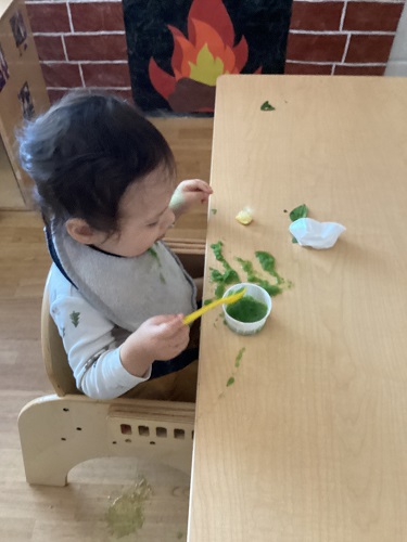 An infant sitting at the table using a spoon to scoop green juice mixture