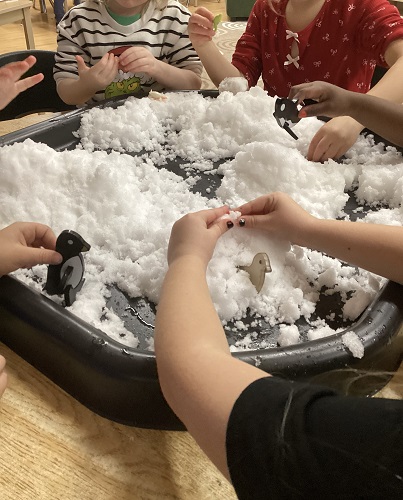 4 School Age 1 children sitting around the tuff tray filled with snow, some are holding magnetic penguins and some are holding orca's, some are manipulating the snow with their fingers