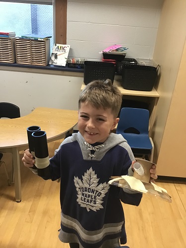 School Age 2 child standing smiling at the camera holding the spaceship and binoculars he created using loose parts