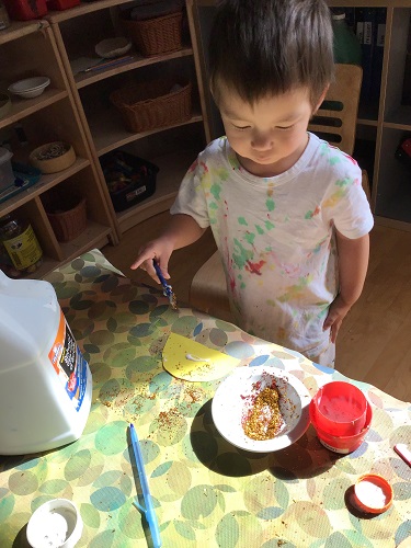 Preschool child standing at the table holding a glue stick looking at the bowl of glitter on the table
