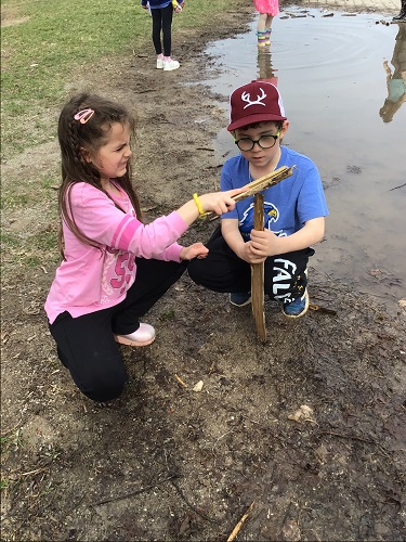 1 school age child crouching down while holding a stick into the ground, another school age child crouching down beside holding a different stick ontop of the one in the ground. There are 3 school age children in the background standing around a large puddle