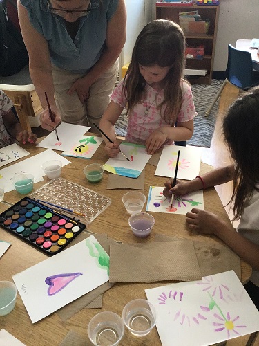 3 children sitting at a table with paper and paintbrushes painting on their paper with the educator standing and showing them a step by step