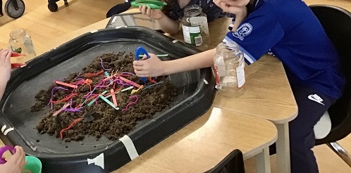 3 School age children sitting around the tuff tray holding tweezers with mud and shredded paper on the table