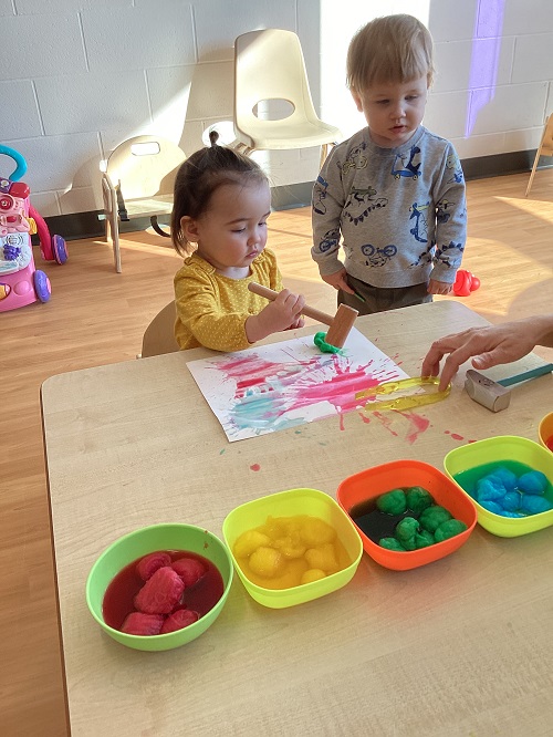 An infant child using a wooden hammer and colourful cotton balls to create a Holi inspired art piece
