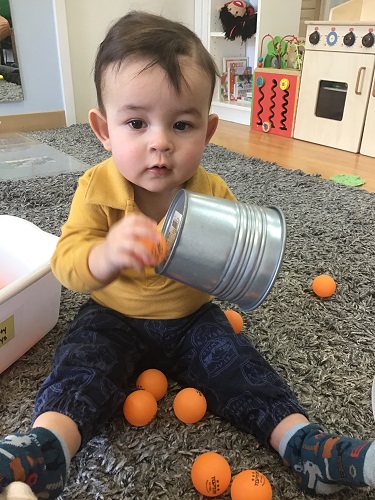An infant is holding a metal bucket and baning a ping pong on the bottom of the bucket.