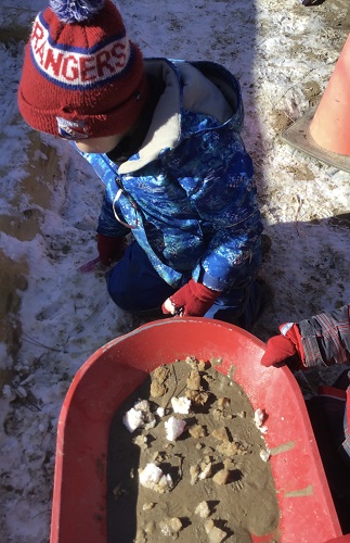 A preschooler has a wheelbarrow that they have put mud and snow in.