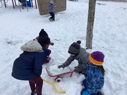 School-age children patting snow down in wheelbarrow