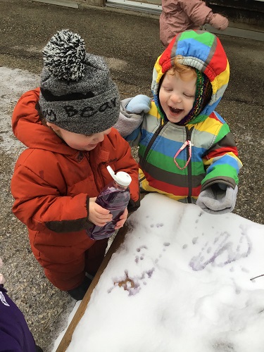 Two toddlers are standing by some snow getting ready to squirt coloured water onto the snow.
