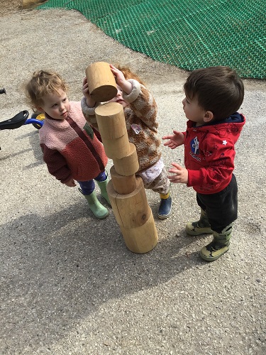 Two toddlers are watching another toddler build a tower with some blocks.