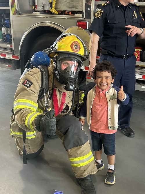 A child giving a thumbs up while standing with a firefighter.