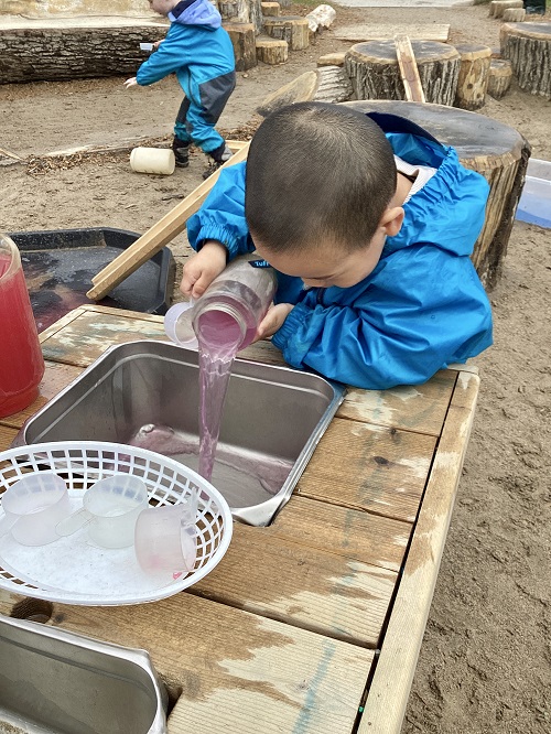 A child pouring a container of water into the mud kitchen sink.