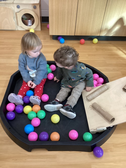 2 toddler children sitting in a tuff tray at the bottom of the ball ramp surrounded by balls