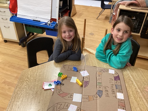 2 School Age 1 children sitting at the table with a home made board game on the table in front of them, both School Age children are smiling at the camera
