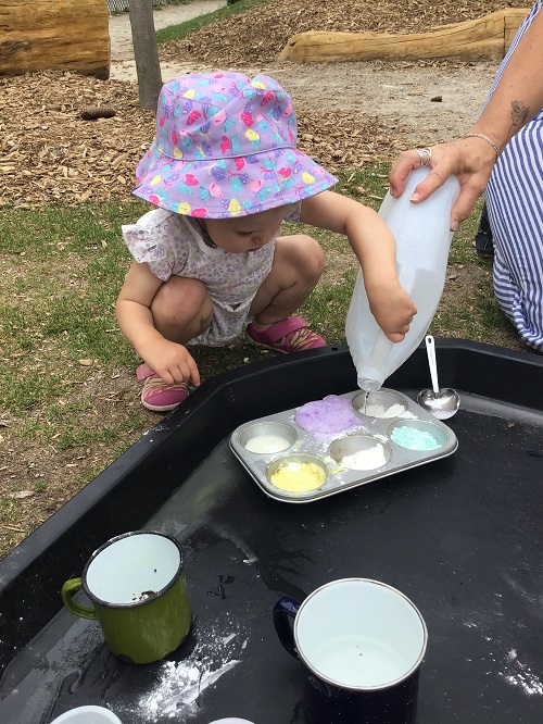Infant crouched bside black tray, in tray is a muffin pan with blue, pink, yellow pastel paint