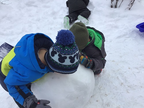 Two children placing their cheeks on a large snowball.