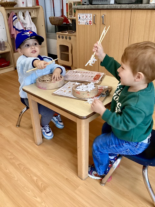 2 preschool children sitting at a table engaging in imaginative play with play noodles, bowls and chopsticks