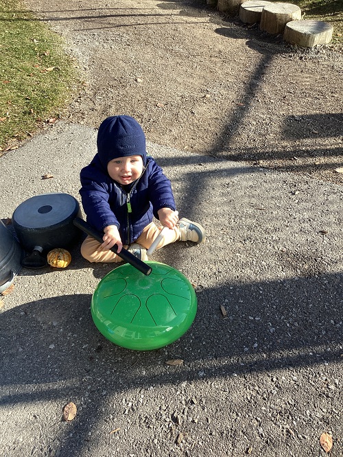 An infant using a metal drum and wooden drumstick outside
