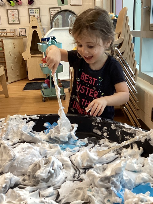 A preschool child excitedly using a spoon to scoop shaving cream into a bowl