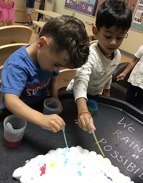 2 boys standing at black tray painting white cotton ball cloud making a rainbow